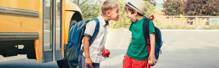 Two funny happy Caucasian boys kids showing tongues each other near yellow bus on 1 September day. Education back to school. Children learn and study. Web banner header for website.の写真素材