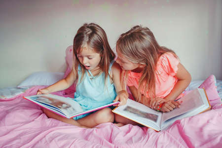 Happy Caucasian sisters girls reading books in bedroom. Children siblings at home spending time together. Kids education development learning. Real people authentic lifestyle.の写真素材