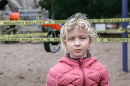 Portrait of sad upset Caucasian girl on closed playground outdoor. Kids play area locked with yellow...