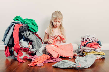Mommy little helper. Cute Caucasian girl sorting clothes. Adorable child arranging organazing clothing. Kid with messy stack of clothes things on floor. Home chores housework for children.の写真素材