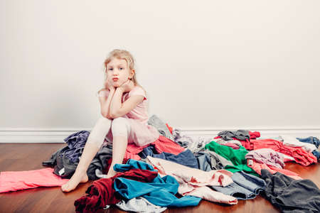 Mommy little helper. Bored Caucasian girl sorting clothes. Tired funny child arranging organazing clothing. Kid with messy stack of clothes things on floor. Home chores housework.の写真素材