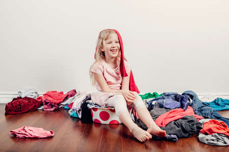 Mommy little helper. Cute Caucasian girl sorting clothes. Adorable funny child arranging organazing clothing. Kid with messy stack of clothes things on floor. Home chores housework for children.の写真素材