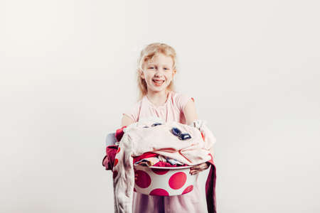 Mommy little helper. Smiling Caucasian girl child holding wash-basin with clothes and laundry detergent pods. Kid with messy stack of clothes. Home chores housework for children.の写真素材