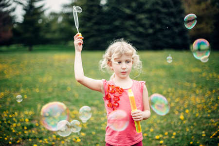 Preschool Caucasian blonde girl blowing soap bubbles in park on summer day. Child having fun...