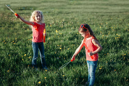 Preschool Caucasian girls blowing soap bubbles in park on summer day. Kids having fun outdoor. Authentic happy childhood magic moment. Lifestyle seasonal activity for children.の写真素材