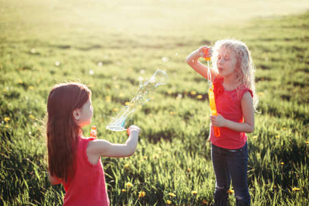 Preschool Caucasian girls blowing soap bubbles in park on summer day. Kids having fun outdoor. Authentic happy childhood magic moment. Lifestyle seasonal activity for children.の写真素材