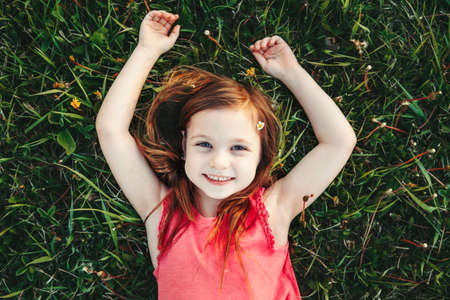 Cute smiling Caucasian girl resting in grass on meadow. Child lying on ground. Outdoor fun summer children activity. Kid having fun outside. Happy childhood lifestyle. View from top above.の写真素材