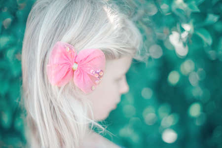 Portrait of little blonde Caucasian girl with pink hair bow. Pretty pensive sad child kid. Girly girl with red hair clip looking away. Lonely child. Soft selective focus. Blurry background. の写真素材