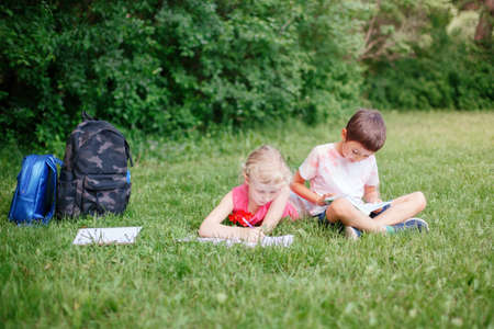 Young Caucasian school girl and boy friends doing school homework in park outdoor. Children kids reading book and writing with pencil. Children education learning studying together.の写真素材