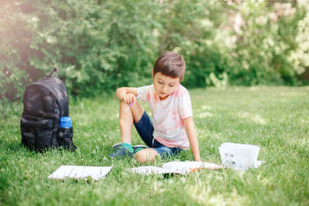 Young Caucasian school boy sitting in park outdoor doing school homework. Child kid writing in notebook with pencil outside. Self education learning studying. Early development for children.の写真素材