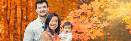 Asian Chinese mother and Caucasian father dad with baby girl in autumn fall park. Family together outdoor in nature. Seasonal holiday and authentic lifestyle. Banner header for website.の写真素材