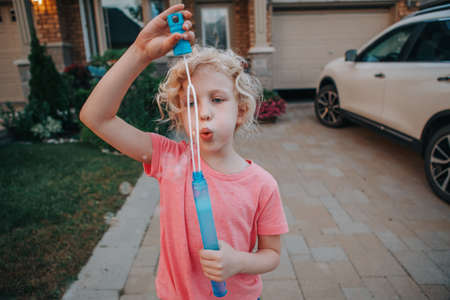 Young Caucasian girl blowing soap bubbles on home front yard. Child having fun outdoor on sunset. Authentic happy childhood magic moment. Lifestyle seasonal activity for children.の写真素材