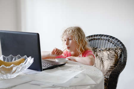 Young girl eating food and watching cartoons on laptop. Child with digital gadget during meal. Kid using modern technology entertainment while eating snack. New generation childhood concept.の写真素材