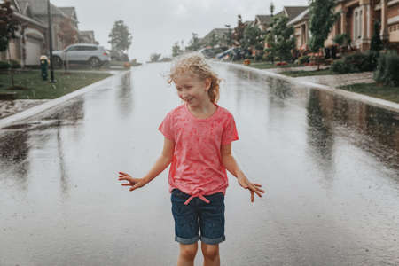 Cute adorable girl running splashing under rain on street road. Child having fun during rain shower storm. Seasonal summer outdoor activity for kids. Freedom and happy childhood lifestyle. の写真素材