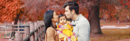 Asian Chinese mother and Caucasian father dad holding kissing baby girl wrapped in blanket. Family in autumn fall park outdoor. Seasonal holiday Thanksgiving. Web banner header for website.の写真素材
