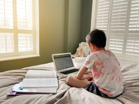 Caucasian boy child sitting in bed and learning online on laptop Internet. Virtual class lesson on...