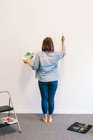 Lifestyle creative hobby and freelance artistic work side job concept. Caucasian woman artist hand painting murals on walls indoor at apartment or studio school with acrylic paints.の写真素材