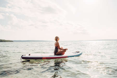 Middle age Caucasian woman practicing yoga on paddle sup surfboard at sunset. Female stretching...
