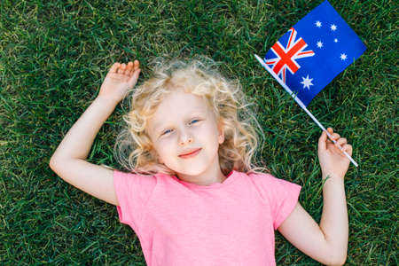 Adorable cute happy Caucasian girl holding Australian flag. Smiling child lying on grass in park holding Australia flag. Kid citizen celebrating Australia Day holiday in January outdoor.の写真素材