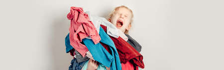Mommy little helper. Adorable funny tired child arranging organazing clothing. Kid holding messy stack pile of clothes things. Home chores housework. Web banner header for website.の写真素材