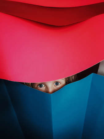 Funny kid playing at home. Child girl sitting in homemade paper house. Creativity and early development for kids. Baby boy girl eyes looking through paper. Abstract geometry. Hide and seek game.の写真素材