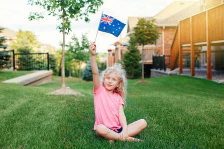 Adorable cute happy Caucasian girl holding Australian flag. Smiling child sitting on grass in park holding Australia flag. Kid citizen celebrating Australia Day holiday in January outdoor.の写真素材