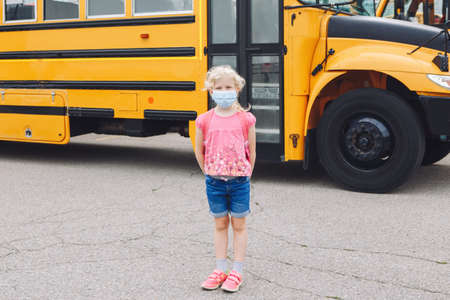 Happy Caucasian girl student wearing face mask near yellow bus. Kid with personal protective...