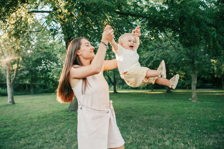 Young mother spinning toddler baby boy outdoor. Parent playing with child son in park. Authentic lifestyle funny moment. Happy family life concept. Childhood and motherhood.の写真素材
