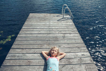 Dreams and relaxation. Girl child with closed eyes lying on wooden lake dock on sunny day. Kid dreaming enjoying summer outdoor. Happy childhood lifestyle. View from above overhead.の写真素材