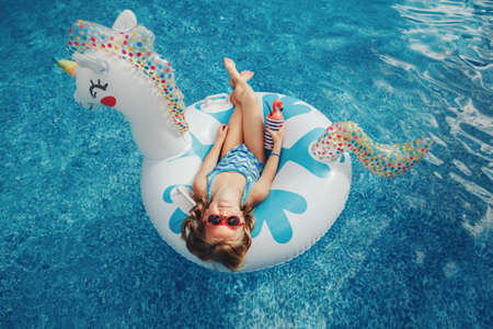 Cute adorable girl in sunglasses with drink lying on inflatable ring unicorn. Kid child enjoying...