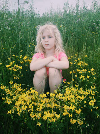 Portrait of little cute Caucasian blonde girl sitting in grass and yellow flowers on meadow. Pensive bored tired kid thinking about future. Child enjoying summer day outdoor.の写真素材