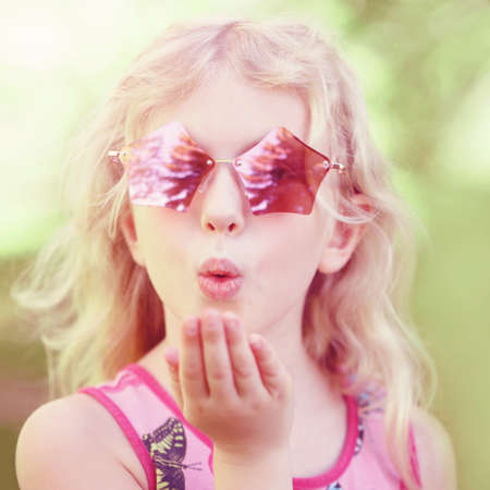 Young girl posing in fancy pink pentagonal shaped sunglasses outdoor. Cute adorable stylish Caucasian child with long blonde hair sending air kiss. Cool hipster kid having fun.の写真素材
