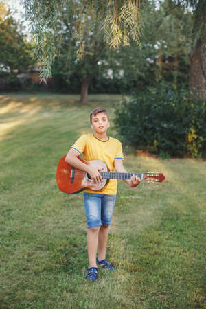 Hard of hearing preteen boy playing guitar outdoor. Child with hearing aids in ears playing music and singing song in park. Hobby art activity for children kids. Authentic childhood moment.の写真素材
