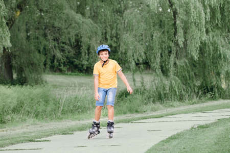 Happy smiling Caucasian preteen boy in helmet riding roller skates on road in park on summer day....