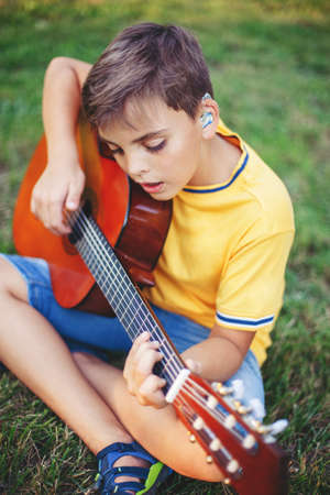 Hard of hearing preteen boy playing guitar outdoor. Child with hearing aids in ears playing music and singing song in park. Hobby art activity for children kids. Authentic childhood moment.の写真素材