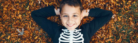 Halloween holiday. Happy child boy in skeleton costume lying on pile of yellow leaves outdoor. Smiling boy preparing for Halloween themed event. Autumn fall fun. Web banner header.の写真素材