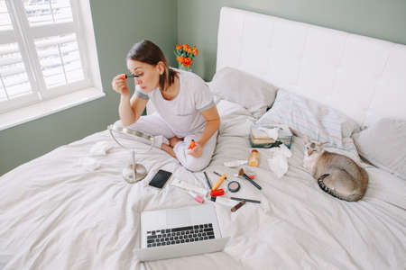 Middle age Caucasian woman applying her makeup in morning. Young woman sitting in bed at home bedroom doing her morning routine in front of laptop. Real authentic lifestyle indoor.の写真素材