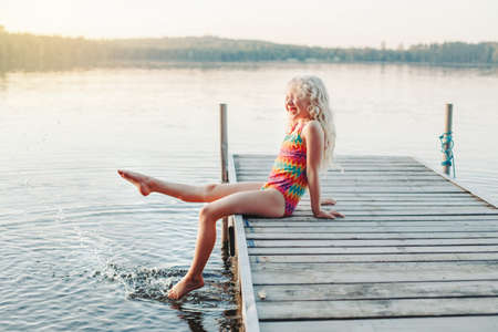 Funny happy cute Caucasian blonde girl child sitting on wooden deck pier by lake. Smiling laughing kid in swimsuit splashing with legs in water. Summer fun outdoor activity. Happy childhood lifestyle.の写真素材