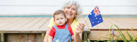 Old woman grandmother with grandson baby holding Australian flag. Old lady with kid child on backyard waving Australia flag. Family citizens celebrating Australia Day. Web banner header.の写真素材
