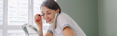 Middle age Caucasian woman applying makeup in morning and talking on phone. Young woman doing her...