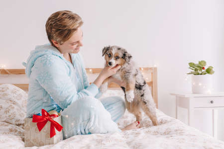 Young happy Caucasian woman playing with cute puppy dog pet at home. Pet owner celebrating Christmas holiday alone. Holiday celebration together with furry little friend.の写真素材