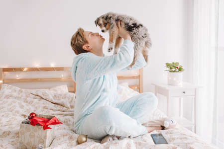 Young happy Caucasian woman playing kissing cute puppy dog pet at home. Pet owner celebrating Christmas holiday alone. Holiday celebration together with furry little friend. Funny holiday gift.の写真素材