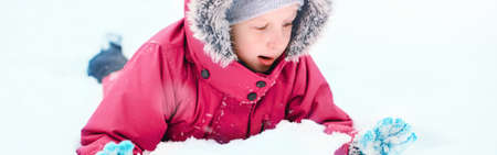 Funny Caucasian girl in winter clothes playing with snow. Cute child lying on ground during cold winter snowy day at snowfall. Kids outdoor seasonal activity. Web banner header.の写真素材