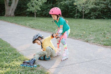 Girl helping boy to stand up on roller blades after falling. Caucasian friends boy and girl riding on roller skates in park on summer day. Seasonal outdoor children activity sport. Kids summer sport.の写真素材