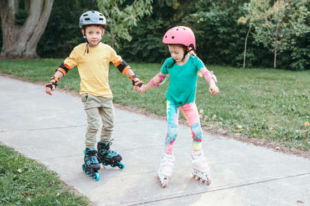 Happy Caucasian friends boy and girl in helmets riding on roller skates in park on summer day....