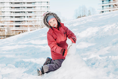 Funny happy girl child in warm clothes playing with snow in park outside. Child kid making snowman during cold winter sunny bright day. Kids outdoor seasonal activity.の写真素材