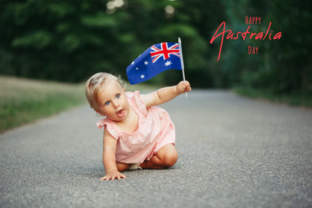 Happy Australia Day. Holiday card with text. Cute adorable Caucasian baby girl waving Australian flag. Funny child crawling on street road in park celebrating Australia Day holiday outdoor.の写真素材