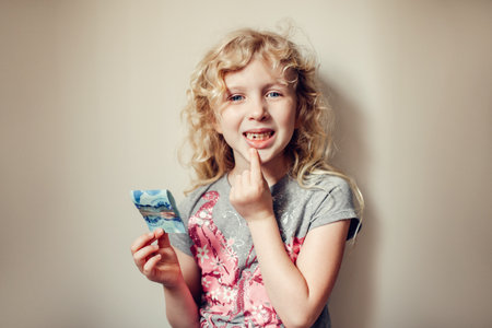 Caucasian blonde girl showing her missing tooth in mouth and holding money from tooth fairy. Proud...