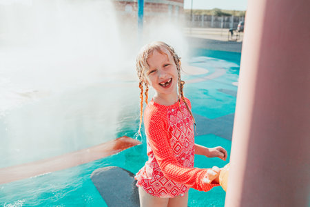 Cute adorable laughing Caucasian funny girl playing on splash pad playground outdoor on summer day. Smiling child having fun in water. Seasonal water sport recreational activity for kids.の写真素材