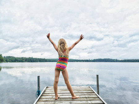 Blonde girl in swimsuit standing on wooden lake river dock with her hands raised up. Harmony with nature and happy healthy active childhood. Summer outdoor water activity. View from back.の写真素材